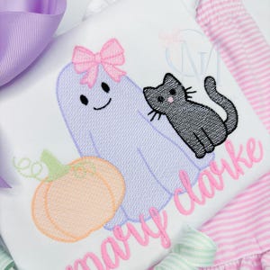 May include: A white shirt featuring an embroidered light purple ghost with a pink bow, a black cat, and an orange pumpkin. The name Mary Clarke is embroidered in pink cursive script below the designs. The shirt is paired with pink striped fabric.