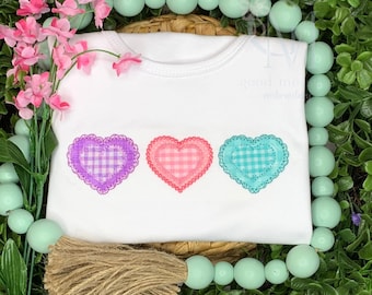 May include: White t-shirt with three embroidered hearts. The hearts are pink, purple, and blue with a white and pink gingham pattern.
