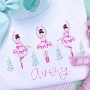May include: White t-shirt embroidered with three ballerinas in pink tutus and pointe shoes, with green Christmas trees. The name "Avery" is embroidered in pink script below the ballerinas. An aqua bow is in the background.
