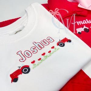 May include: White long-sleeved shirt with the name "Joshua" embroidered in red and light blue. The shirt features red tractors and hearts embroidered along a green grassy line. A red shirt is visible in the background.
