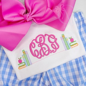 May include: A white shirt with a pink monogram and a stack of colorful books with an apple on top. The shirt is paired with blue and white gingham shorts.