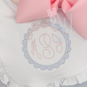 May include: White baby bib with a pink and blue embroidered monogram. The bib features a scalloped edge and a large pink bow at the top. The monogram includes the letters "M" and "G".