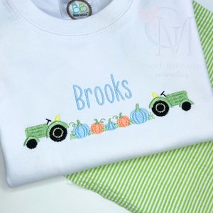 May include: White baby onesie with blue embroidered text "Brooks" and a green tractor pulling a line of pumpkins. The pumpkins are blue, orange, and green.