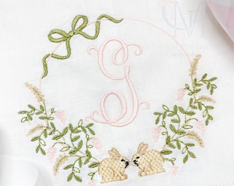 May include: Embroidered white linen tea towel with a green bow, a pink floral wreath, and two bunnies kissing inside the wreath. The letter G is embroidered in pink in the center of the wreath.