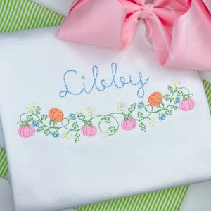 May include: White garment with embroidered floral design featuring pink pumpkins, orange gourds, blue flowers, and white daisies. The name "Libbey" is embroidered in blue script above the floral design. A pink bow is in the background.