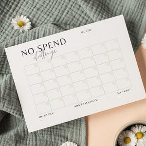 Op de afbeelding: Een witte No Spend Challenge-planner met een raster voor het bijhouden van uitgaven. De planner bevat secties voor de maand, niet-essentiële zaken en de 'waarom' van de gebruiker. Madeliefjes en een groene doek staan op de achtergrond.