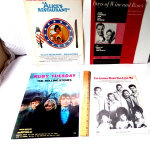 May include: A collection of vintage sheet music and songbooks. Titles include "Alice's Restaurant," "Days of Wine and Roses," "Ruby Tuesday," and "I'm Gonna Make You Love Me." A wooden ruler is also present.