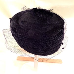 May include: A dark navy blue hat with a delicate net veil. The hat has a rounded crown and a small bow detail. The veil is attached around the brim and drapes down, adding a touch of elegance. A vintage-style accessory.