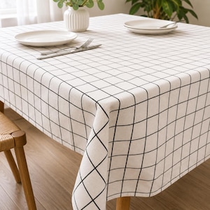 May include: A white tablecloth with a black grid pattern on a wooden table. Two white plates, cutlery, and a small vase with greenery are on the table. The tablecloth has a simple, modern design.