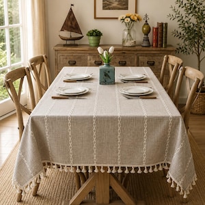 May include: A beige rectangular tablecloth with white vertical embroidered stripes and a tassel trim. The table is set with white plates, cutlery, and a blue vase with flowers. Wooden chairs surround the table.