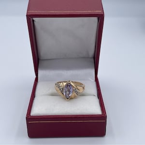 Könnte beinhalten: Ein goldener Ring mit einem hellvioletten, marquiseförmigen Edelstein. Der Ring ist in einem goldenen Band mit kleinen Akzentsteinen auf jeder Seite gefasst. Präsentiert in einer roten Schmuckschatulle mit weißem Satineinsatz.
