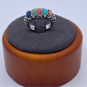 May include: A silver ring featuring five gemstones in blue, green, pink, turquoise, and peach. The ring has a decorative filigree design and a smooth band. The ring is displayed on a grey surface.