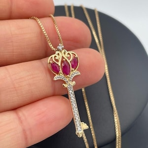 May include: A gold key pendant necklace featuring three marquise-cut red rubies set in the bow of the key, accented with small clear stones along the stem and bow, suspended from a delicate gold box chain with a secure clasp mechanism.