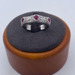 Könnte beinhalten: Ein silberfarbener Ring mit drei rund geschliffenen roten Rubinen in einer strukturierten Ringschiene. Jeder Rubin ist von einem seilartigen Metalldetail und kleinen klaren Steinen umgeben. Die Schiene weist an den Seiten ein eingraviertes Muster auf.