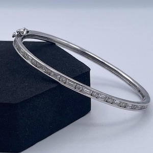 Pode incluir: Uma pulseira rígida de tom prateado com cravação em canal, alternando pedras claras redondas e de corte baguette. A pulseira tem um acabamento metálico polido e inclui um mecanismo de fecho de dobradiça seguro na parte superior.