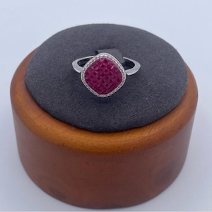 Może przedstawiać: Srebrny pierścionek z kwadratowym, rubinowym kamieniem centralnym. Kamień centralny ma teksturowaną powierzchnię i jest otoczony przez halo z małych, przezroczystych kamieni. Pierścionek jest prezentowany na szarej poduszce na drewnianym stojaku.