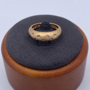 Può includere: Un anello d'oro con finitura opaca, incastonato con piccole pietre preziose trasparenti. L'anello è esposto su un cuscino grigio scuro all'interno di un portagioie in legno. Il design dell'anello presenta gemme distanziate uniformemente.