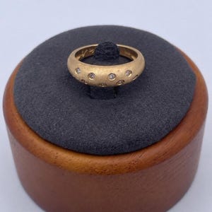 Può includere: Un anello d'oro con finitura opaca, incastonato con piccole gemme chiare e uniformemente distanziate. L'anello è esposto su una superficie grigio scuro, che si trova su una base di legno.
