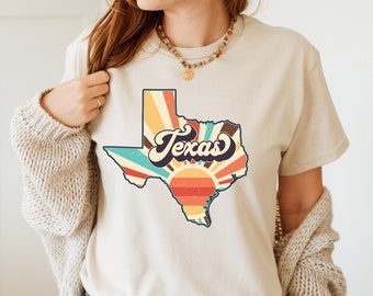 May include: A beige t-shirt with a vintage-style graphic of the state of Texas with a sunburst design and the word "Texas" in a retro font.