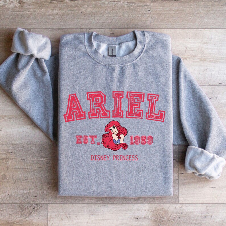 May include: A grey sweatshirt featuring the name "ARIEL" in red, block lettering. Below the name is a graphic of Ariel, the Disney princess, with the text "EST. 1989" and "DISNEY PRINCESS" in red.