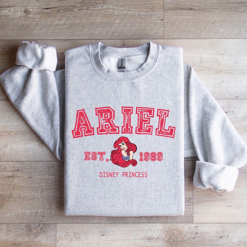 May include: A light grey sweatshirt with the name "ARIEL" in red block letters, with "EST. 1989" and a graphic of Ariel from The Little Mermaid. The words "DISNEY PRINCESS" are printed below the graphic.
