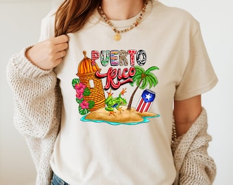 May include: A beige t-shirt with a colorful graphic design featuring a cartoon fort, palm tree, frog, and a can with the Puerto Rican flag. The text "PUERTO RICO" is written in a colorful, stylized font.