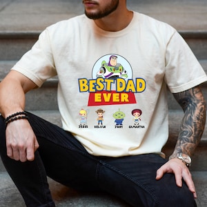 K&ouml;nnte beinhalten: Cremefarbenes T-Shirt mit dem Schriftzug "BEST DAD EVER" in Gelb und Rot, mit einem Cartoon-Bild von Buzz Lightyear. Darunter befinden sich Cartoon-Figuren namens Jessi, Kelvin, Tony und Samantha. Das Shirt ist ein Freizeitartikel.