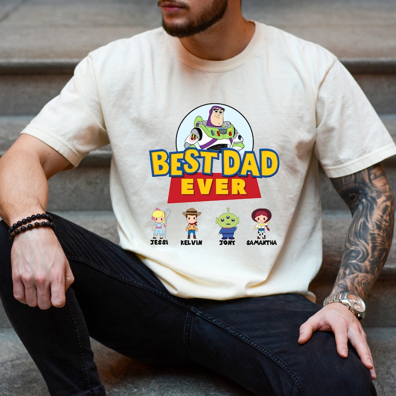 K&ouml;nnte beinhalten: Cremefarbenes T-Shirt mit dem Schriftzug "BEST DAD EVER" in Gelb und Rot, mit einem Cartoon-Bild von Buzz Lightyear. Darunter befinden sich Cartoon-Figuren namens Jessi, Kelvin, Tony und Samantha. Das Shirt ist ein Freizeitartikel.