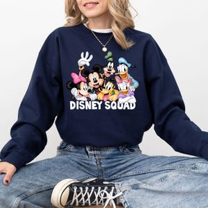 Puede incluir: Sudadera azul marino con un gr&aacute;fico de personajes de Disney, incluyendo Mickey Mouse, Minnie Mouse, Donald Duck y Goofy. El texto "DISNEY SQUAD" est&aacute; impreso en blanco. La sudadera la lleva una persona.