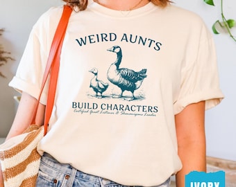 May include: An ivory t-shirt featuring a dark teal graphic of a goose and a gosling. The text reads WEIRD AUNTS BUILD CHARACTERS Certified Great Listener & Shenanigans Leader. The shirt has short sleeves and a crew neckline, shown in a casual style.