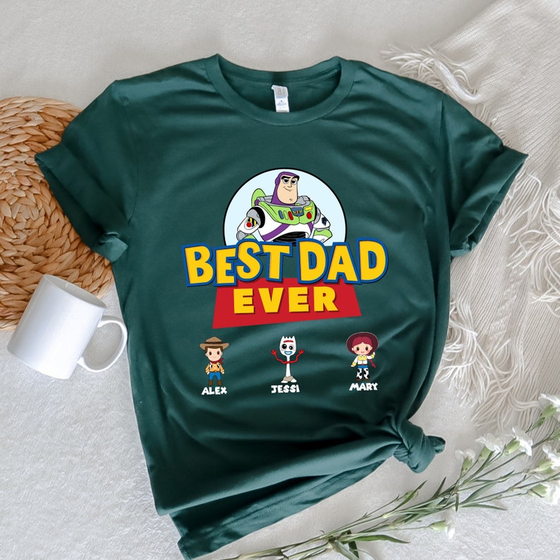 K&ouml;nnte beinhalten: Dunkelgr&uuml;nes T-Shirt mit einer Grafik von Buzz Lightyear und dem Text "BEST DAD EVER" in Gelb und Rot. Unter dem Text befinden sich Cartoon-Illustrationen von Woody, Forky und Jessie mit den Namen Alex, Jessi und Mary. Links ist eine wei&szlig;e Tasse zu sehen.