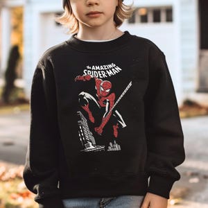 Puede incluir: Sudadera negra con un dise&ntilde;o de Spider-Man en una pose din&aacute;mica, con el texto "The Amazing Spider-Man" en blanco. El dise&ntilde;o incluye un paisaje urbano. La sudadera es de cuello redondo.