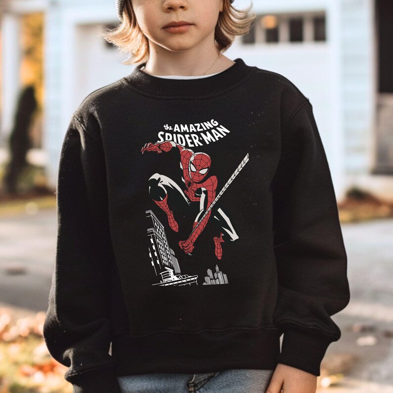 Puede incluir: Sudadera negra con un dise&ntilde;o de Spider-Man en una pose din&aacute;mica, con el texto "The Amazing Spider-Man" en blanco. El dise&ntilde;o incluye un paisaje urbano. La sudadera es de cuello redondo.