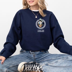 Op de afbeelding: Marineblauw sweatshirt met een cartoon eend gezicht ontwerp in een geruite cirkel. Het woord "NAME" staat eronder gedrukt. Het sweatshirt is effen marineblauw van kleur. De persoon draagt een spijkerbroek en sneakers.