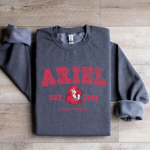 May include: A charcoal grey sweatshirt featuring the name "ARIEL" in red, block lettering. Below the name are the words "EST. 1989" and a graphic of Ariel from Disney's The Little Mermaid. The words "DISNEY PRINCESS" are below the graphic.