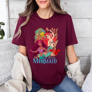 May include: A maroon t-shirt featuring a graphic print of Ariel, Flounder, and Sebastian from The Little Mermaid. The text "The Little Mermaid" is printed below the characters.