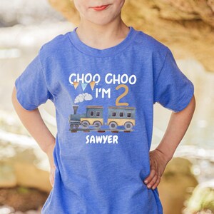 May include: A blue short-sleeve t-shirt featuring a graphic of a train with the text "CHOO CHOO I'M 2" and the name "SAWYER" printed below. The design includes a large yellow number two, colorful bunting flags, and a small cloud of steam.