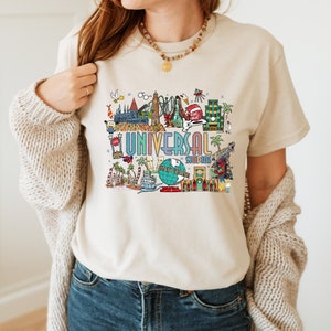 May include: A beige t-shirt with a colorful graphic design featuring various attractions from Universal Studios. The design includes the Universal Studios logo, iconic rides, and characters like the Minions and the Hogwarts castle.