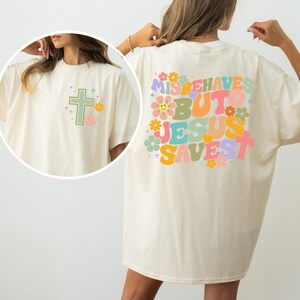 May include: Cream-colored t-shirt with a cross and floral design on the front and the words "MISBEHAVES BUT JESUS SAVES" on the back. The graphic is in pastel colors.