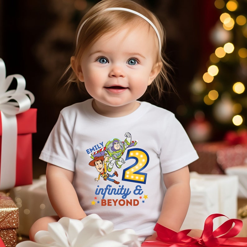 May include: A white short-sleeved shirt with a colorful graphic of Woody and Buzz Lightyear from Toy Story. The graphic includes the number "2" with lights around it and the text "Emily infinity & BEYOND".