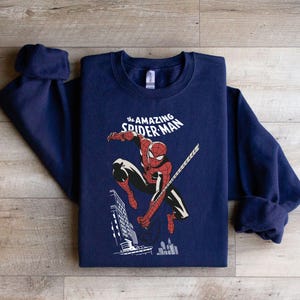 Puede incluir: Una sudadera azul marino con un gr&aacute;fico de Spider-Man en una pose din&aacute;mica, con el texto "the AMAZING SPIDER-MAN" arriba. La ilustraci&oacute;n muestra a Spider-Man con su cl&aacute;sico traje rojo y negro, con un tel&oacute;n de fondo de la ciudad.