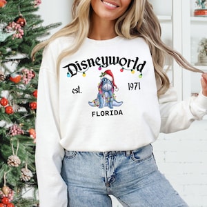 May include: White sweatshirt with the text "Disneyworld est. 1971 FLORIDA" in black script. The design includes a blue donkey wearing a Santa hat and Christmas lights. The sweatshirt is long-sleeved.