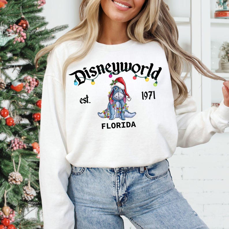 May include: White sweatshirt with the text "Disneyworld est. 1971 FLORIDA" in black script. The design includes a blue donkey wearing a Santa hat and Christmas lights. The sweatshirt is long-sleeved.