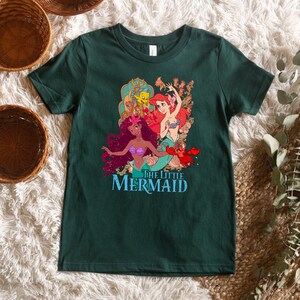 May include: A dark green t-shirt featuring a graphic print of Ariel, Ursula, and Flounder from The Little Mermaid. The text "The Little Mermaid" is printed below the characters.