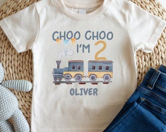May include: A cream-colored short-sleeve t-shirt featuring a graphic of a blue and yellow train with the text "CHOO CHOO I'M 2" and the name "OLIVER" printed below. The design includes small colorful bunting flags and a white cloud.
