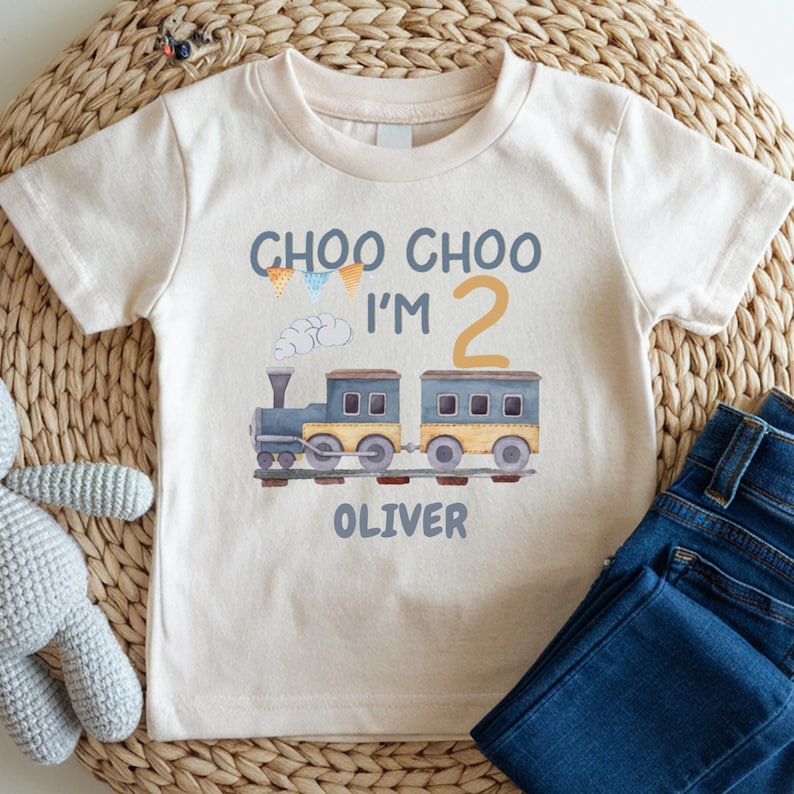 May include: A cream-colored short-sleeve t-shirt featuring a graphic of a blue and yellow train with the text "CHOO CHOO I'M 2" and the name "OLIVER" printed below. The design includes small colorful bunting flags and a white cloud.