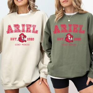 May include: Two sweatshirts, one cream and one khaki green, each displaying the name "ARIEL" in red block letters. Below the name is "EST. 1989" and an image of Ariel, the Disney princess. The words "DISNEY PRINCESS" are printed below the image.