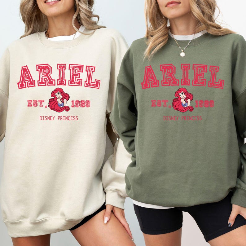 May include: Two sweatshirts, one cream and one khaki green, each displaying the name "ARIEL" in red block letters. Below the name is "EST. 1989" and an image of Ariel, the Disney princess. The words "DISNEY PRINCESS" are printed below the image.