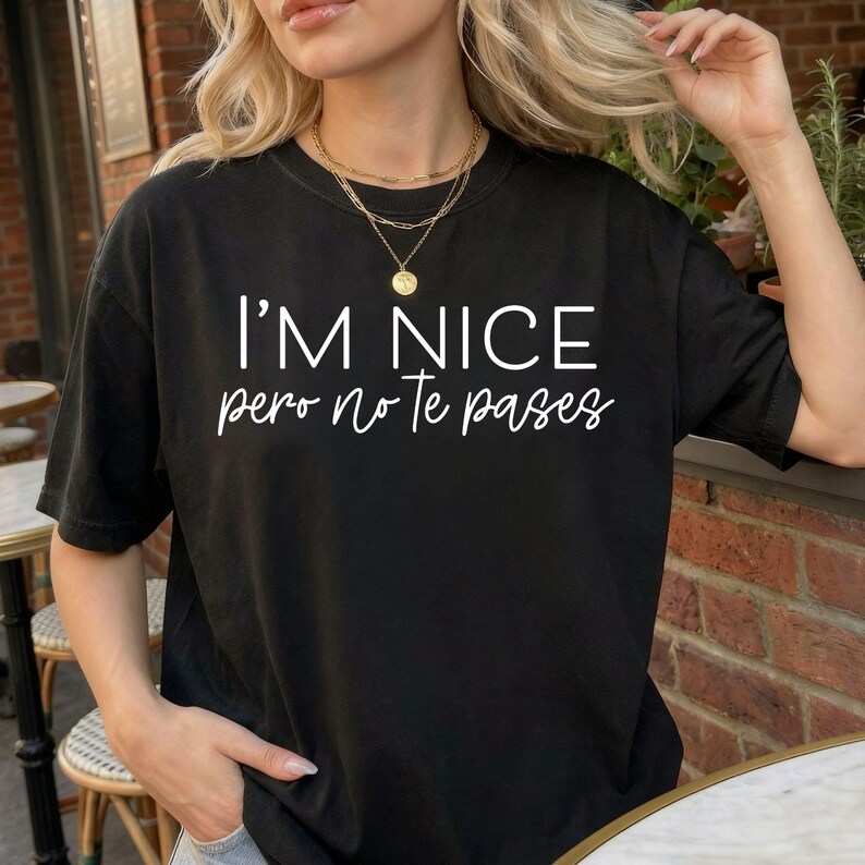 May include: A black short-sleeved t-shirt featuring the white text I'M NICE pero no te pases printed on the front. The shirt has a crew neckline and a relaxed fit, showcasing a combination of bold block lettering and a cursive script font style.