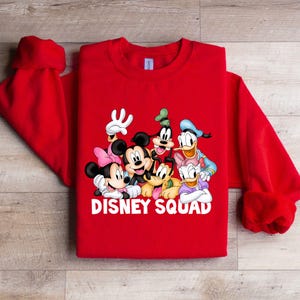 Puede incluir: Una sudadera roja con un gr&aacute;fico de personajes de Disney, incluyendo Mickey Mouse, Minnie Mouse, el Pato Donald y Goofy. El texto "DISNEY SQUAD" est&aacute; impreso en blanco debajo de los personajes. La sudadera tiene cuello redondo y mangas largas.